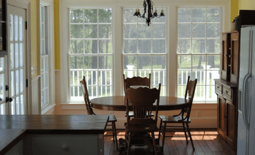Bright kitchen with a round wooden table and chairs, large windows letting in natural light, and a view of greenery outside.