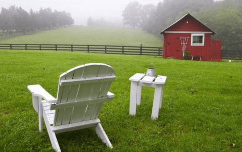 A white Adirondack chair and small table in a grassy field, with a red shed and misty hills in the background.