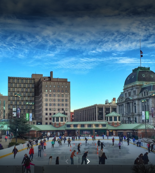 A bustling ice skating rink surrounded by buildings under a blue sky, with people enjoying skating activities.