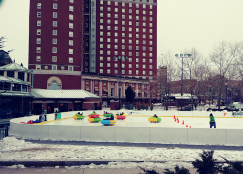 A winter scene with people on a snow-covered ice rink, surrounded by a white barrier and a tall red building in the background.