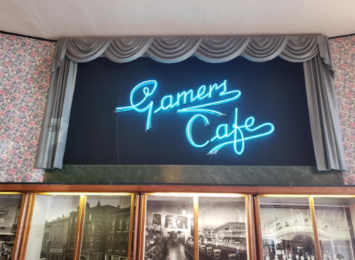 Neon sign reading "Gamer Cafe" against a floral wallpaper backdrop with decorative curtains.