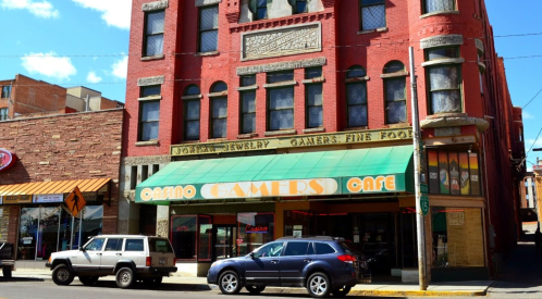 Historic red brick building with "Gamers Cafe" sign, featuring a green awning and parked cars in front.