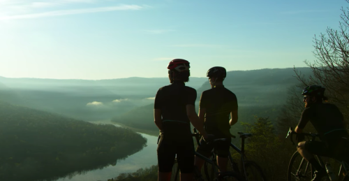 Three cyclists overlook a scenic valley and river at sunrise, surrounded by misty mountains.