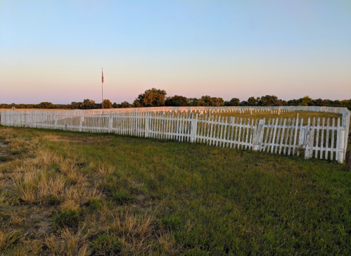 A white picket fence stretches across a grassy field, with a flagpole and trees in the background under a pastel sky.