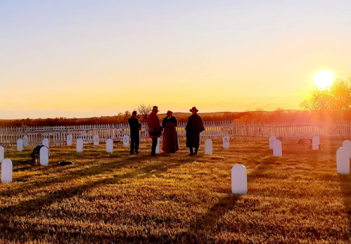 Four people in historical attire stand among white gravestones at sunset, with a warm glow in the background.