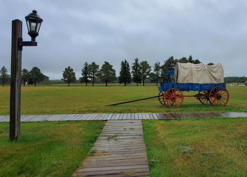 A blue covered wagon on a grassy field, with a wooden path and a lamp post nearby, under a cloudy sky.