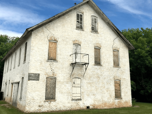 Abandoned two-story building with boarded windows and cracked white brick exterior, set against a blue sky.