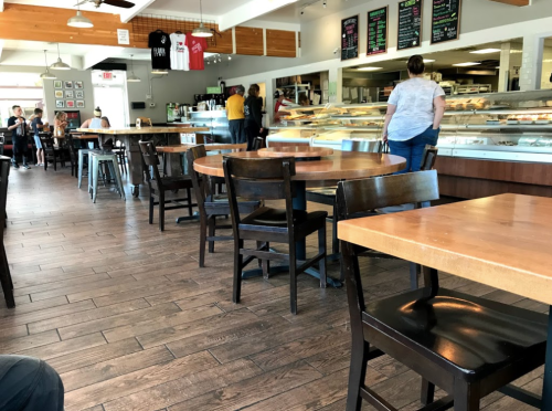 A cozy café interior with wooden tables and chairs, featuring a display counter and customers in the background.