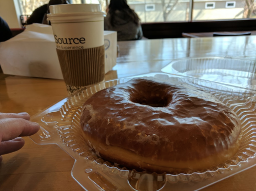 A large glazed donut on a table next to a coffee cup, with a blurred background of a cafe setting.