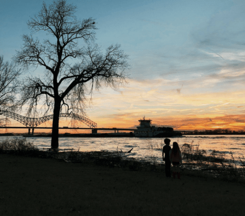 Two children stand by a river at sunset, with a bridge and a tree silhouetted against the colorful sky.