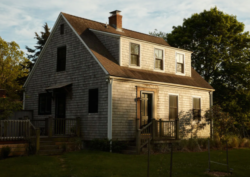 A charming two-story house with wooden shingles, a front porch, and surrounded by greenery under a clear sky.
