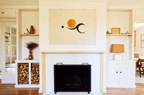 A cozy living room featuring a white fireplace, wooden shelves, and a wall hanging with a sun and abstract shapes.