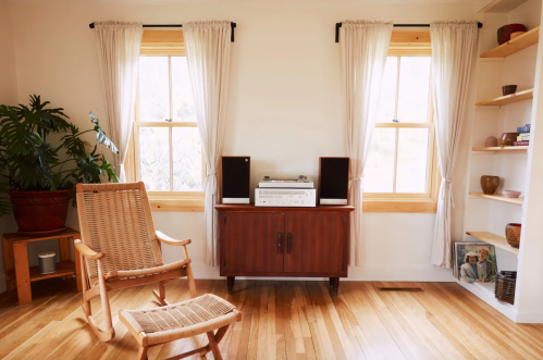 A cozy room with a rocking chair, wooden cabinet, stereo system, and large windows with sheer curtains.