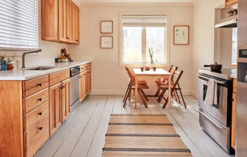 Bright kitchen with wooden cabinets, a dining table, and a woven rug on the floor, featuring natural light from a window.