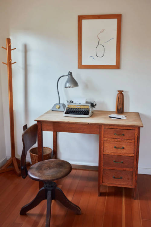 A wooden desk with a typewriter, lamp, and a vase drawing on the wall, accompanied by a chair and coat rack.