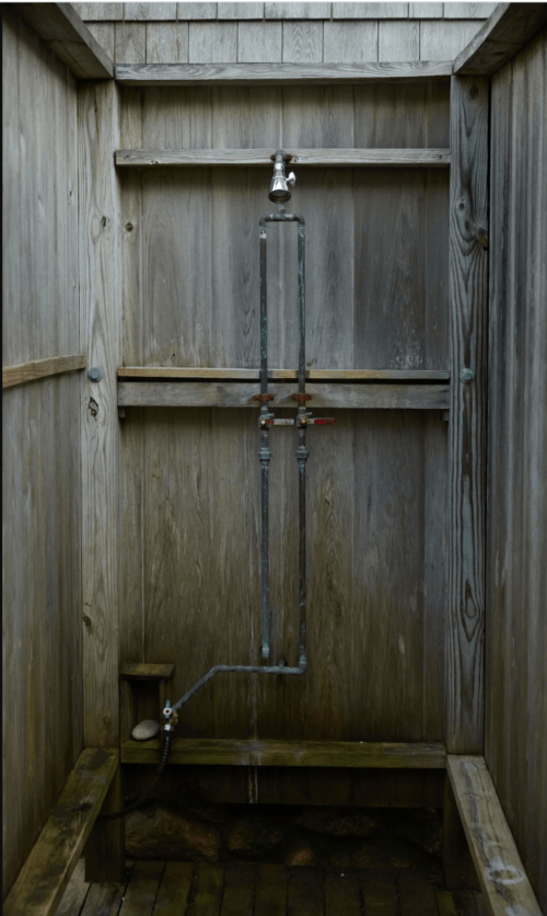 An outdoor shower with wooden walls and a simple metal showerhead and plumbing setup.