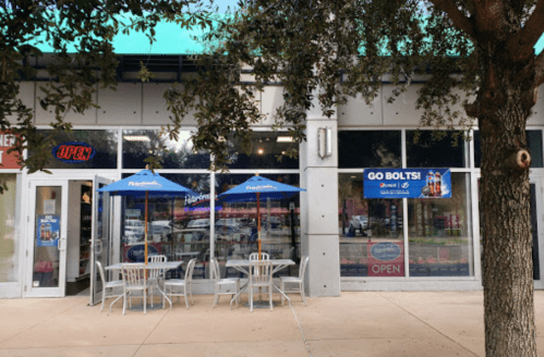 A storefront with an "OPEN" sign, outdoor seating, and banners promoting a sports team. Trees are in the foreground.