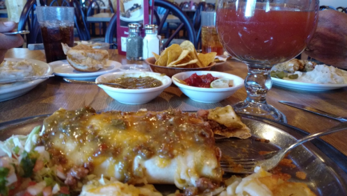 A table set with a plate of enchiladas topped with sauce, sides of salsa, chips, and drinks in a restaurant setting.