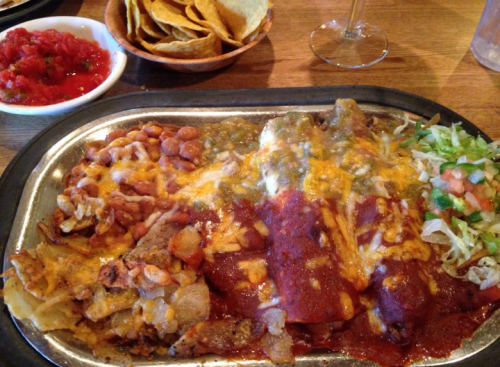 A sizzling plate of enchiladas topped with cheese and sauce, served with beans, rice, salad, and tortilla chips.
