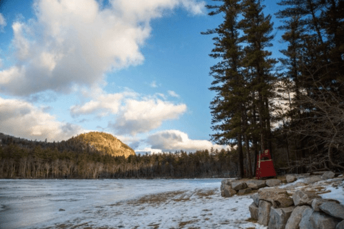 A serene winter landscape featuring a frozen lake, snow-covered ground, tall trees, and a distant rocky hill under a blue sky.
