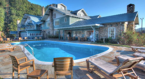 A scenic pool area with lounge chairs, surrounded by a stone building and lush greenery under a clear blue sky.