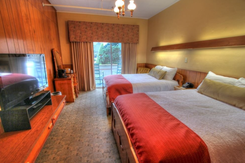 Cozy hotel room with two beds, a TV, and a balcony door leading to an outdoor view. Warm wood accents throughout.