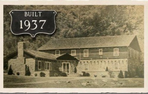 Historic photo of a log cabin-style house with a sign reading "Built 1937" in a wooded area.