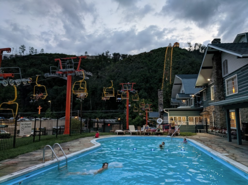 A pool surrounded by mountains, with a chairlift in the background and people swimming under a cloudy sky.