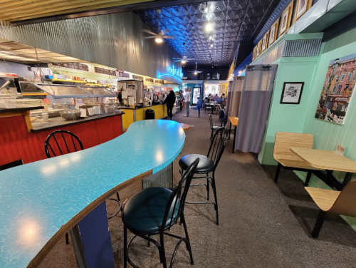 A colorful café interior with a turquoise counter, seating, and a cozy atmosphere, featuring a neon sign and artwork.