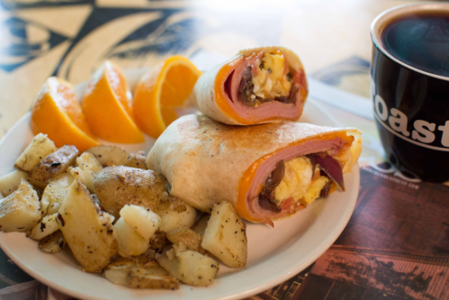 A plate with two breakfast burritos, seasoned potatoes, and orange slices, alongside a cup of coffee.