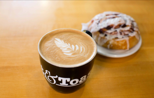 A latte with a leaf design in a black cup next to a cinnamon roll with icing on a wooden table.
