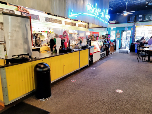 A colorful café interior with a service counter, neon signs, and tables along a carpeted walkway.