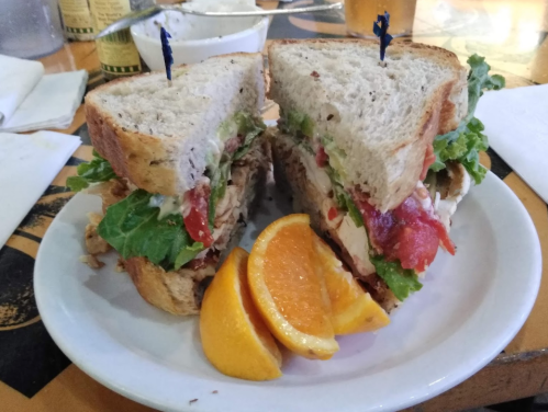 A hearty sandwich with layers of lettuce, tomato, and chicken, served with orange slices on a white plate.