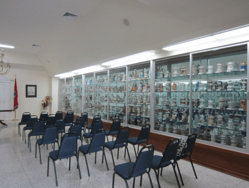 A room with a glass display case filled with various jars, and several blue chairs arranged in front.