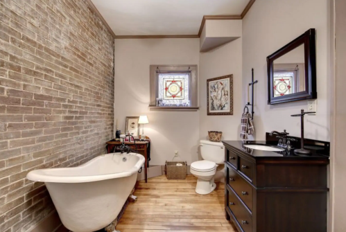 A cozy bathroom featuring a vintage bathtub, wooden flooring, and a brick wall with stained glass windows.