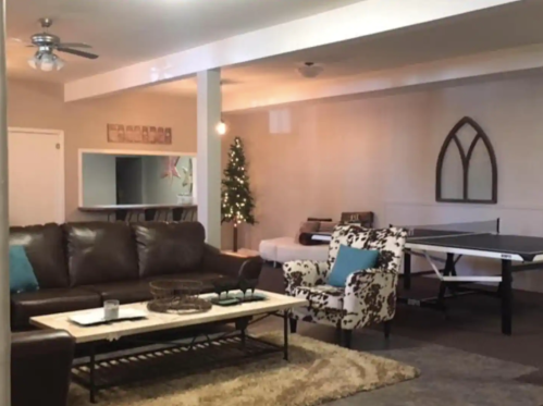 Cozy basement living area with a brown couch, patterned chairs, a ping pong table, and a small Christmas tree.