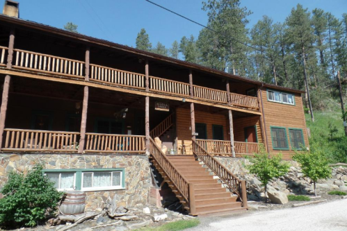A wooden lodge with a large porch, surrounded by trees and a gravel path. Steps lead up to the entrance.