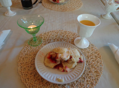 A plate of strawberry shortcake on a table with a green glass candle holder and a white cup of orange juice.