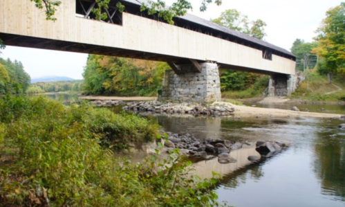 A covered wooden bridge spans a river, surrounded by trees with autumn foliage and rocky riverbanks.