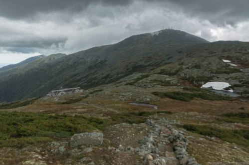 A rocky path leads through a mountainous landscape under cloudy skies, with a building visible in the foreground.