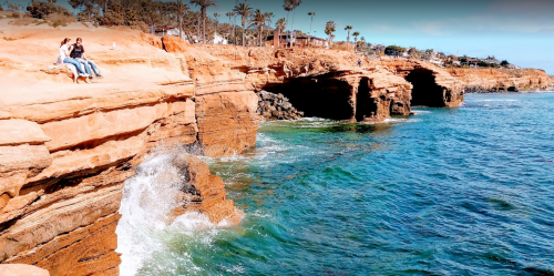 Two people sit on a rocky cliff by the ocean, with waves crashing against the rocks and palm trees in the background.