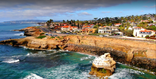 A coastal view featuring cliffs, waves, and colorful houses along the shoreline under a partly cloudy sky.