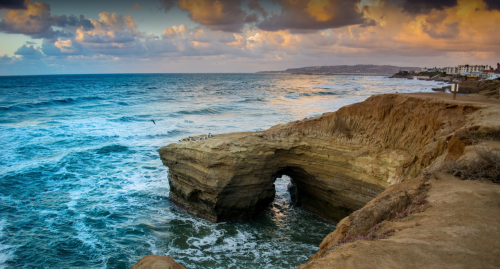 A rocky coastline with an arch, waves crashing below, and a colorful sky at sunset.