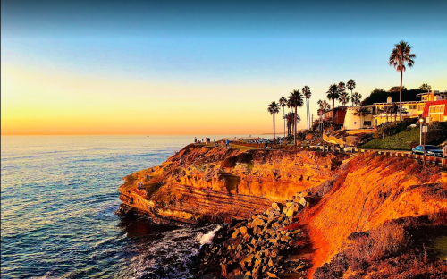 Sunset over a coastal cliff with palm trees, waves crashing below, and colorful buildings along the shoreline.