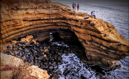 Three people stand on a rocky cliff overlooking the ocean, with waves crashing against the shore below.