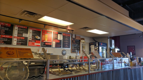 Interior of a restaurant counter displaying a menu with tacos, salads, and drinks, along with food preparation areas.
