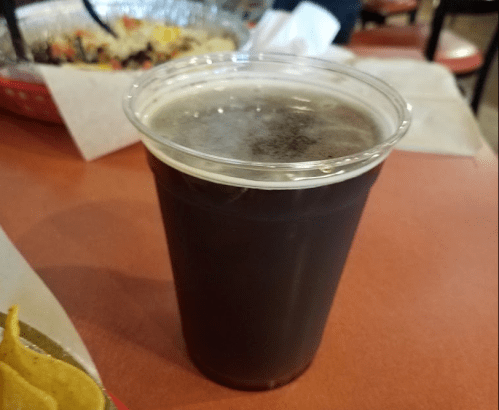 A clear plastic cup filled with dark liquid, placed on a red table, with food items in the background.