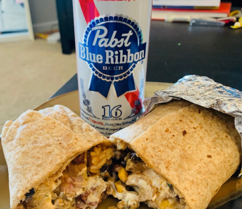 A plate with a cut burrito revealing its filling, next to a can of Pabst Blue Ribbon beer.