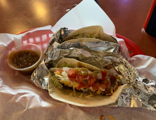 Three tacos wrapped in foil on a red tray, accompanied by a small cup of salsa.