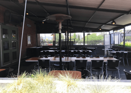 An empty outdoor dining area with tables and chairs under a covered patio, surrounded by greenery.